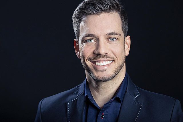A smiling man in a blue shirt and blazer poses against a dark background.