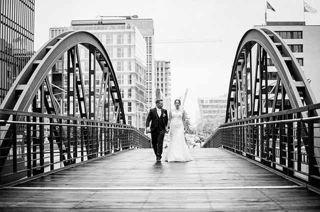 A bride and groom walk hand-in-hand across a modern bridge with a cityscape backdrop in black and white.
