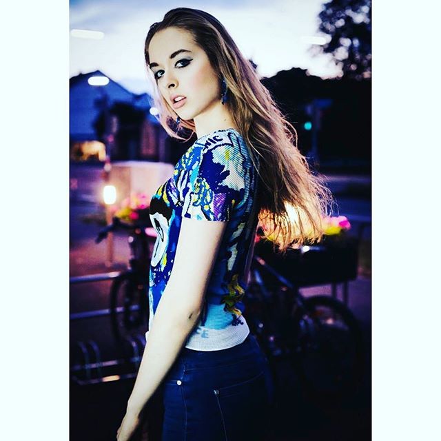A stylish woman poses outdoors at night, wearing a patterned top and jeans, for a fashion or beauty campaign.