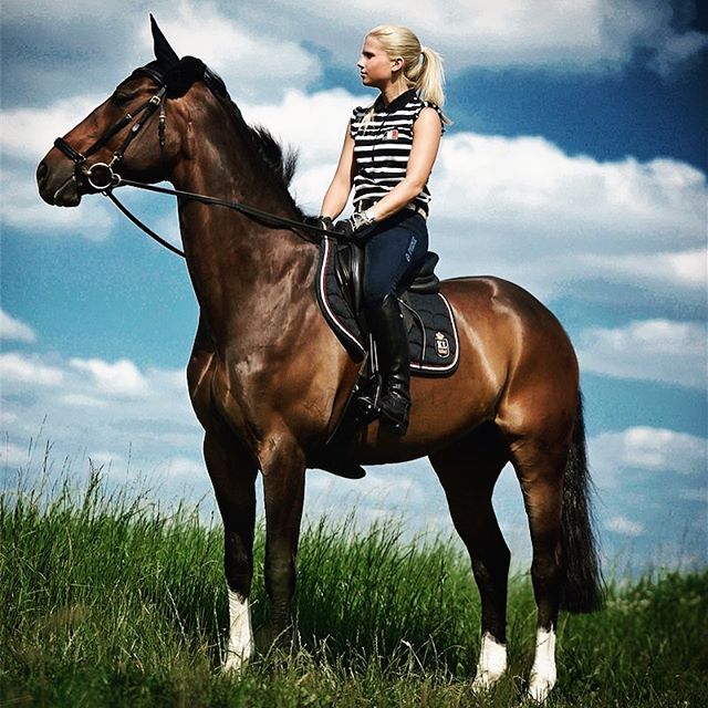 A blonde woman rides a brown horse through a grassy field under a cloudy sky.