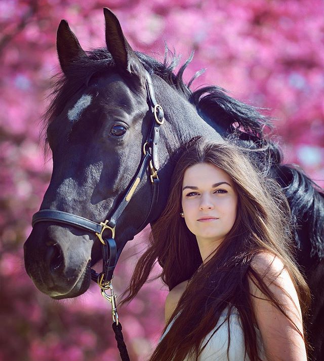 A young woman poses with her black horse in a blooming park setting.