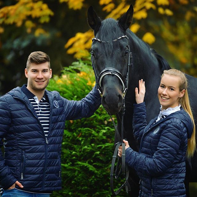 A man and a woman stand with their black horse in a park, smiling and enjoying the scenery.
