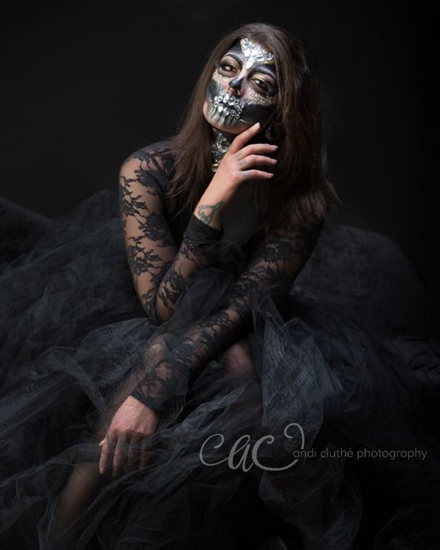 A woman with dramatic skull makeup poses in a black lace dress against a dark backdrop.