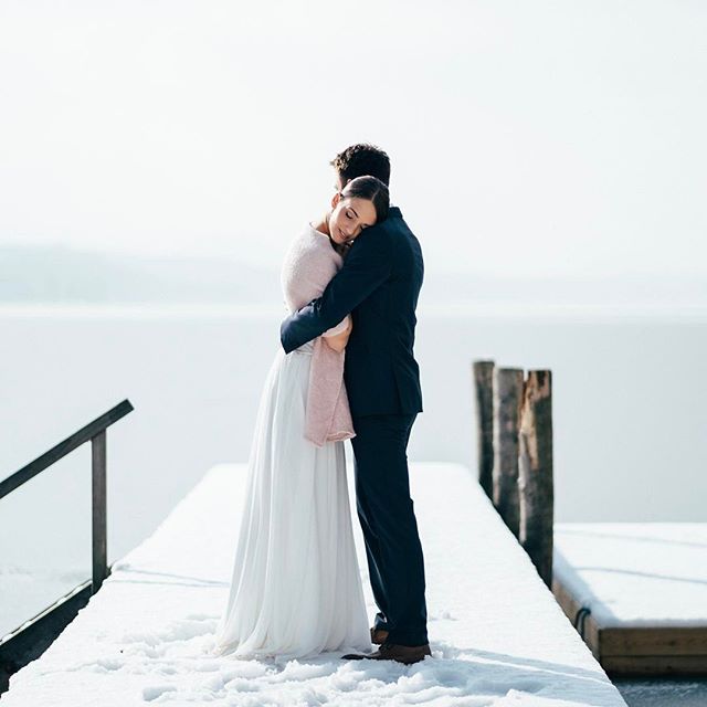 A bride and groom embrace on a snow-covered pier with a serene lake backdrop on their wedding day.