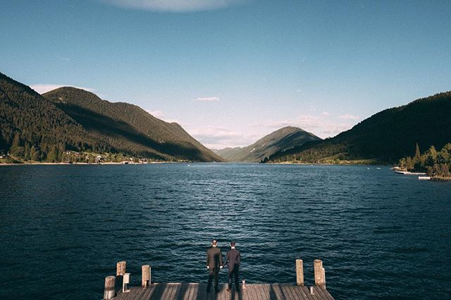 Two men in suits stand on a wooden pier looking out over a calm lake surrounded by mountains.