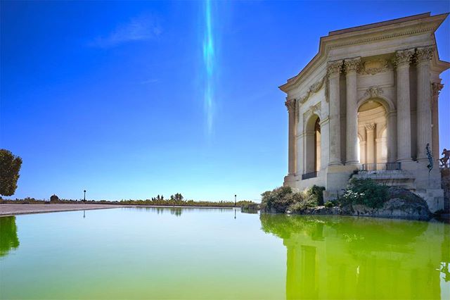 A classical pavilion reflects in a tranquil, green-tinted pool under a clear blue sky. A timeless architectural scene.