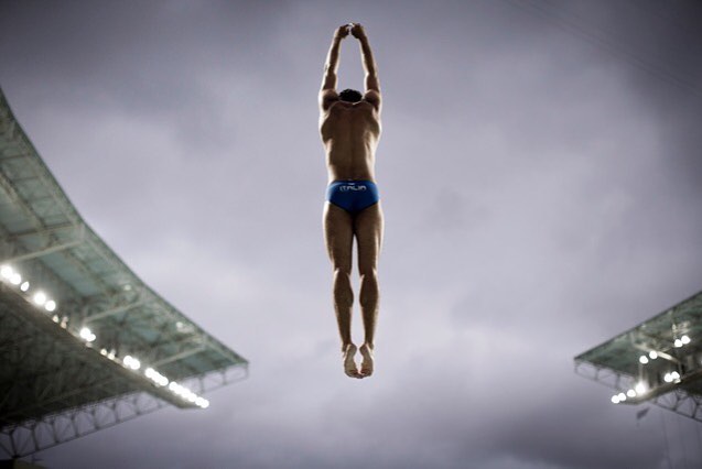 A male diver with arms raised wearing a blue swimsuit prepares to dive from a platform at an outdoor arena.