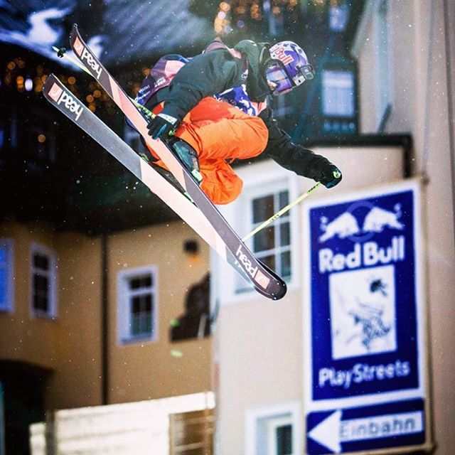 A skier performs a jump in an urban setting with a Red Bull sign nearby.