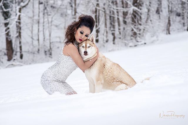 A woman in a dress embraces her husky dog in a serene, snow-covered winter landscape.
