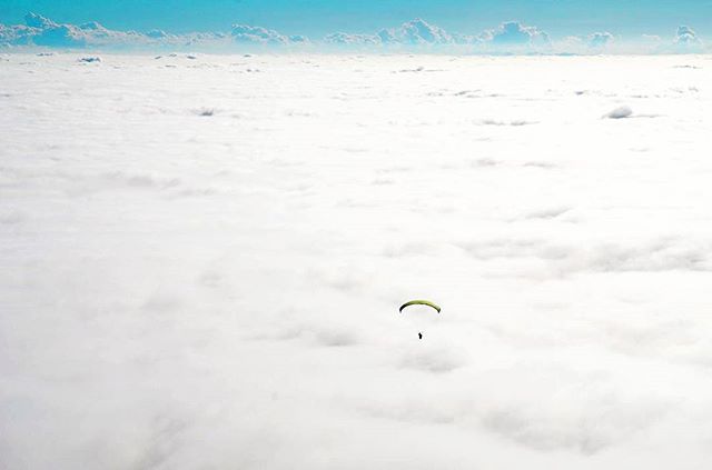 An aerial view shows a person paragliding above soft, fluffy white clouds, against a blue sky.