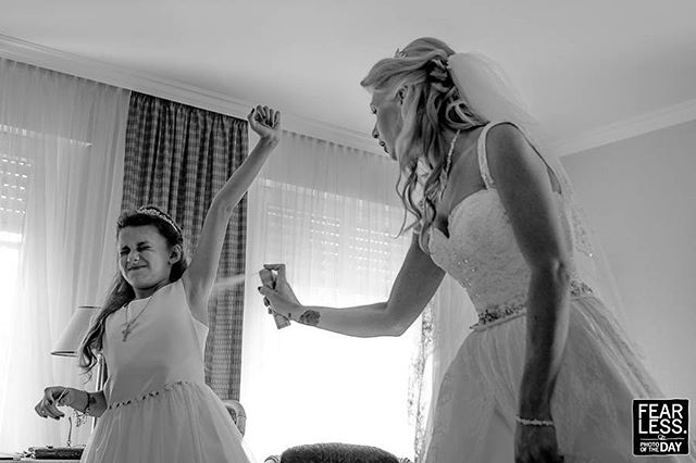 A bride sprays a flower girl with hairspray in a candid black and white wedding photo.