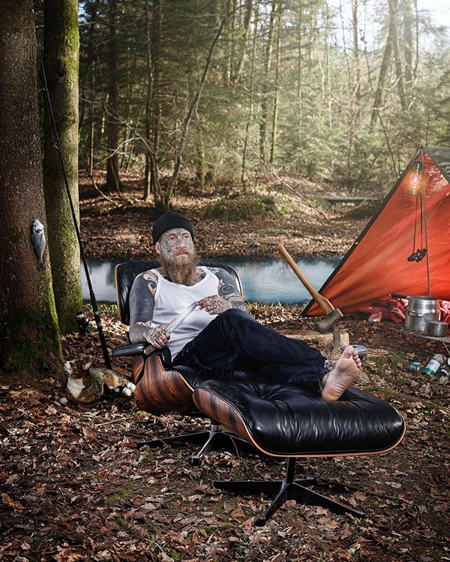 A tattooed man with a beard relaxing in a modern chair in a forest camping scene by a lake.