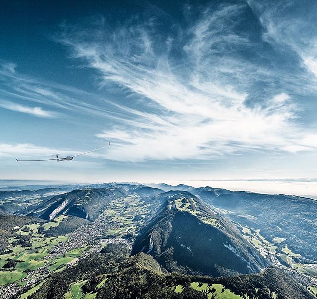 A glider soars above a lush, green mountain valley under a bright blue sky with scattered clouds.