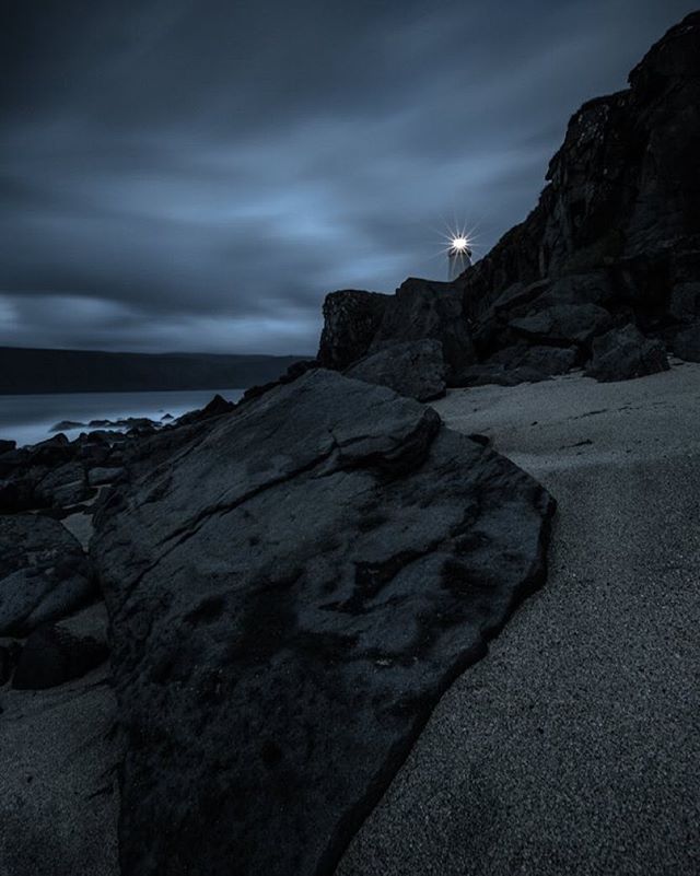 A lighthouse beam shines on a rocky coast at night under a cloudy sky, offering a calm yet mysterious seascape.