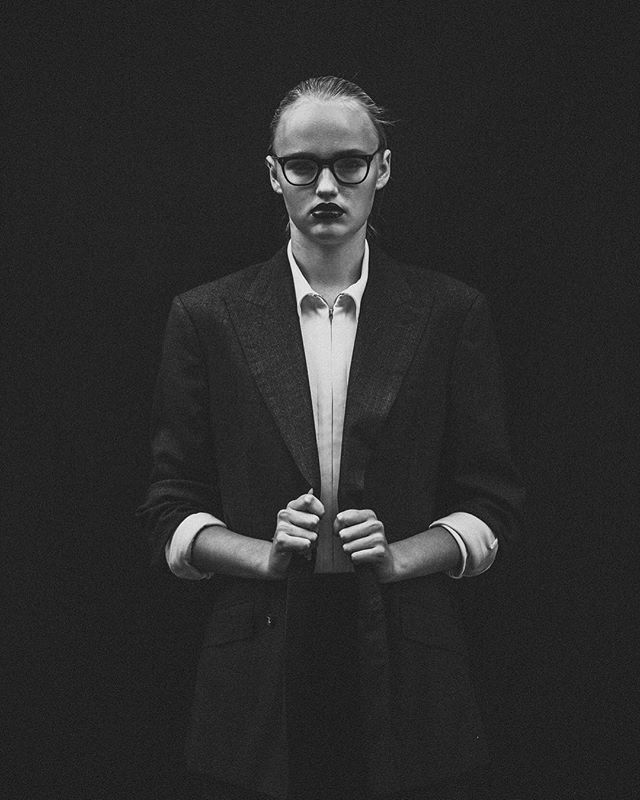 A woman in a suit and glasses poses with a serious expression in a black and white editorial style image.
