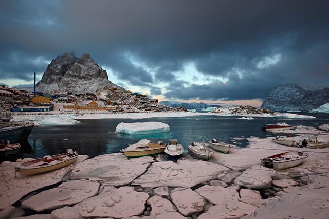 A scenic view of a Greenland fishing village nestled amongst mountains, icebergs, and boats on a cold, overcast day.