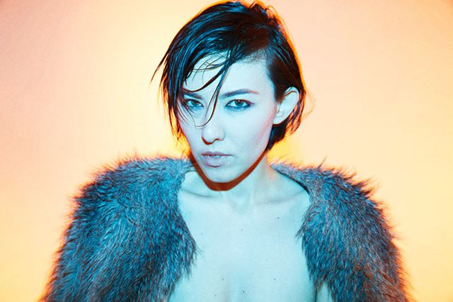 A woman poses in a fur stole under soft, artificial lighting. Editorial shot for fashion advertising.