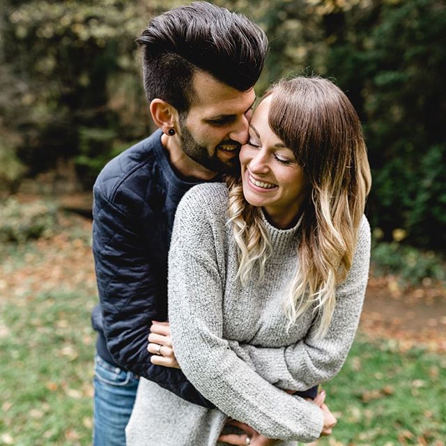 A smiling couple embraces outdoors, sharing a tender moment in a park setting.