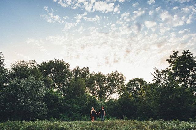 A couple holds hands in a field surrounded by trees under a cloudy sky, creating a natural, intimate moment.