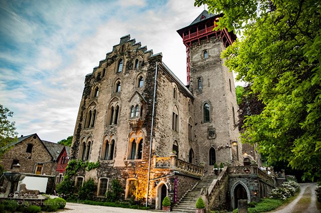 A stone castle with a red-topped tower stands proudly amongst trees under a bright sky.