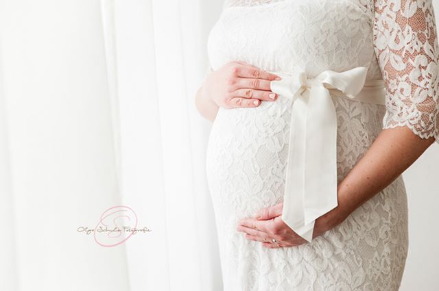 A pregnant woman in a white lace dress holds her belly in a loving, intimate studio portrait.