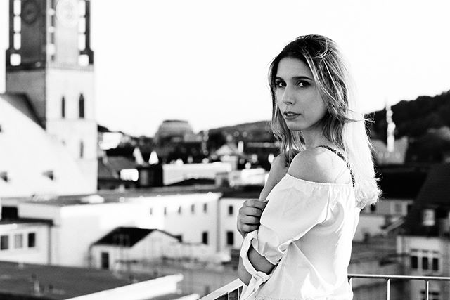 A woman with blonde hair wearing an off-shoulder top poses on a rooftop with a city building backdrop.
