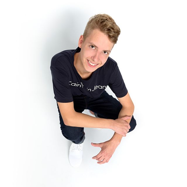 A smiling teenage boy squats and looks up, wearing a t-shirt, jeans, and white shoes on a clean white background.