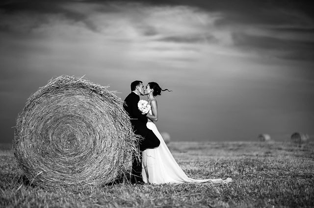 A bride and groom share a kiss next to a hay bale in a black and white wedding photo.