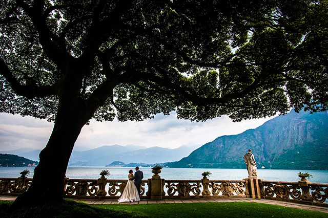 A bride and groom stand on a villa terrace overlooking a lake and mountains on their wedding day.