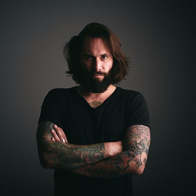 Portrait of a bearded man with tattooed arms, crossing his arms and looking directly at the camera with a serious expression.