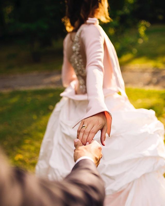A bride leads her groom holding hands, showcasing a wedding ring in a romantic outdoor setting.