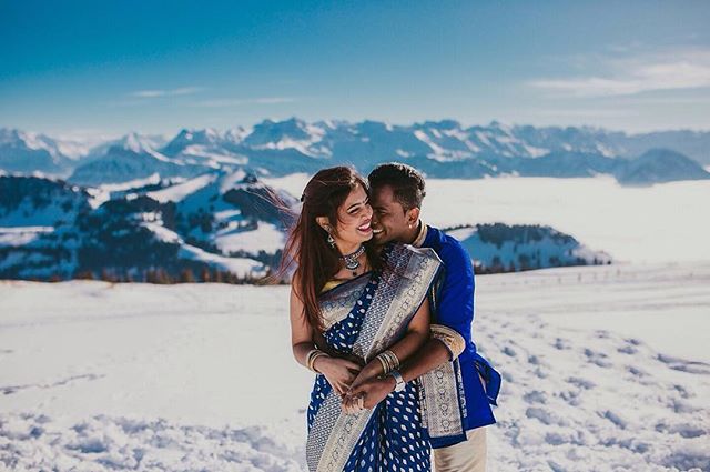 A happy couple in traditional Indian attire embraces in a snowy mountain landscape on a clear, bright day.