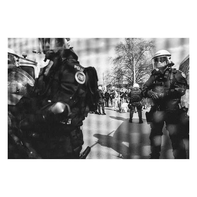 Black and white shot of police officers in riot gear facing a crowd during a protest on a city street.