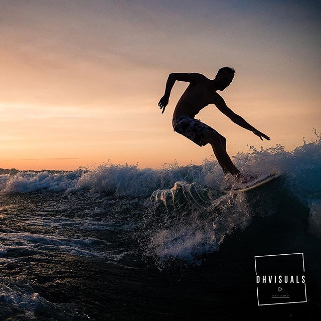 A surfer rides a wave at sunset, silhouetted against the colorful sky.
