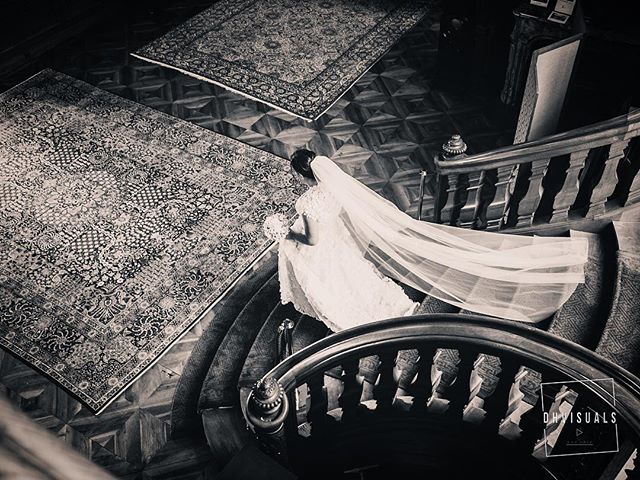 A bride in a white dress descends a staircase in a vintage building. The image is black and white.