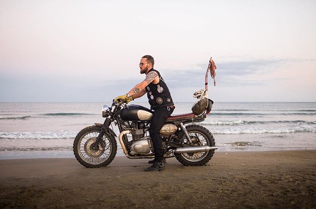 A man with tattoos and sunglasses rides a vintage motorcycle on a sandy beach with the ocean in the background.