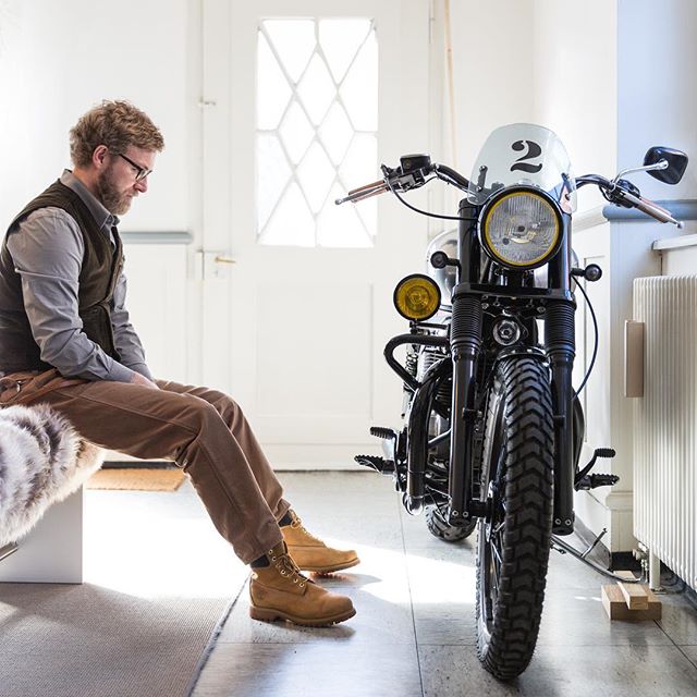 A stylish man sits near a vintage motorcycle in a bright hallway, creating a calm, vintage aesthetic.