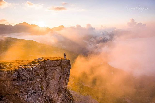 A man stands on a cliff overlooking a mountain range at sunset, creating a scenic and motivational travel landscape.