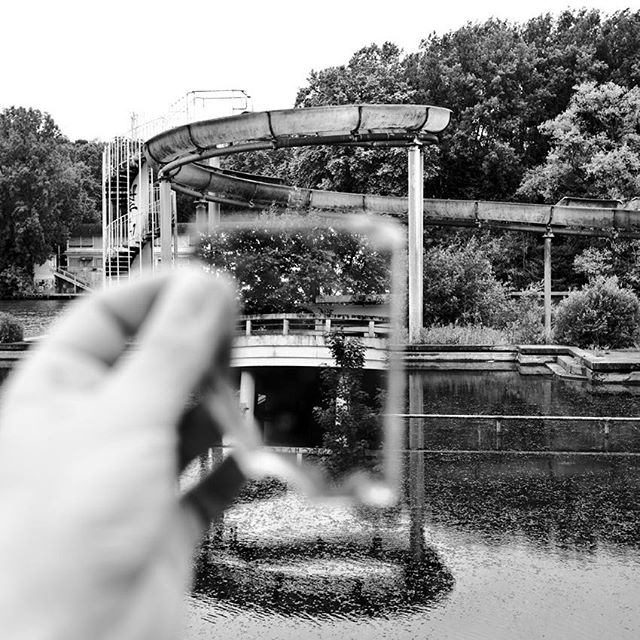 An abandoned water slide sits over a lake, partially visible through a handheld square in a black and white image.