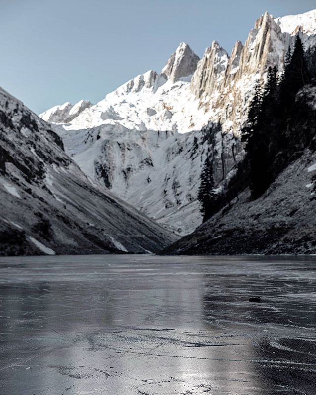A frozen lake sits nestled in a valley, framed by snow-capped mountains under a clear sky.