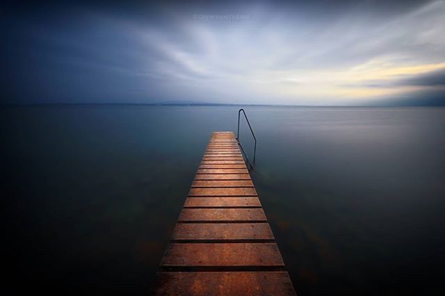 A tranquil lake scene with a wooden pier stretching into the calm water under a soft, cloudy sky.
