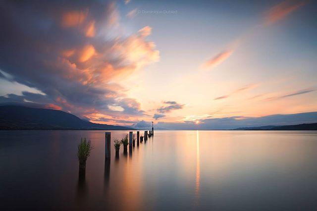 A serene sunset over a tranquil lake with a wooden pier stretching into the calm water beneath a sky painted with pastel clouds.