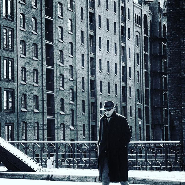 A man in a hat and coat walks on a city street with old warehouse buildings in the background, in black and white.