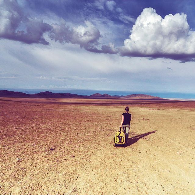 A woman walks across a vast desert with a yellow suitcase, under a cloudy sky, emphasizing adventure and travel.