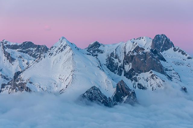 A scenic view of snow-covered mountains with pink-tinged sky and fog at their base.