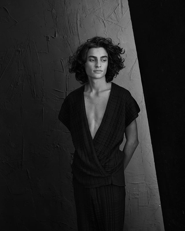 A young man with curly hair poses in a black and white fashion portrait against a textured background.