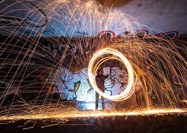 A person spinning steel wool creates light trails against a graffiti-covered wall in an urban tunnel, captured with long exposure.