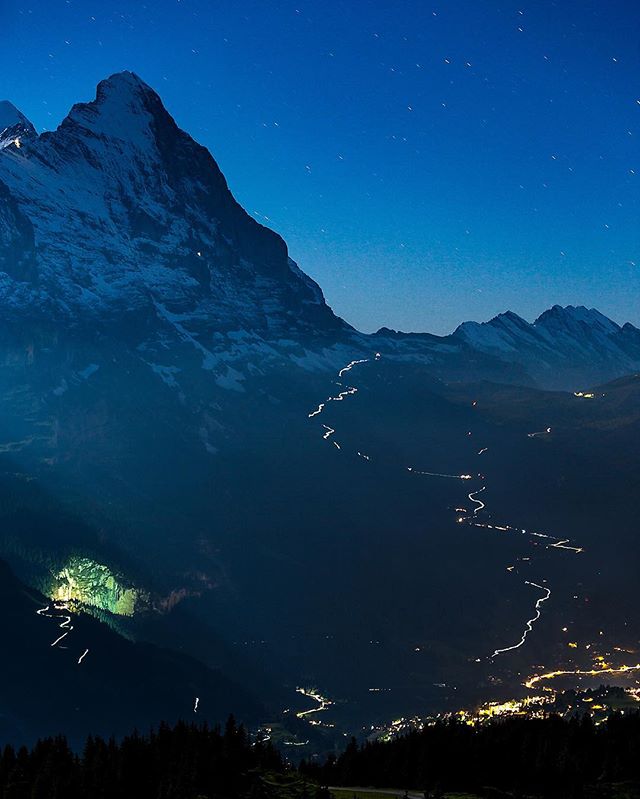 A stunning nightscape capturing snow-capped mountains, a winding trail of city lights, and a serene, tranquil atmosphere.