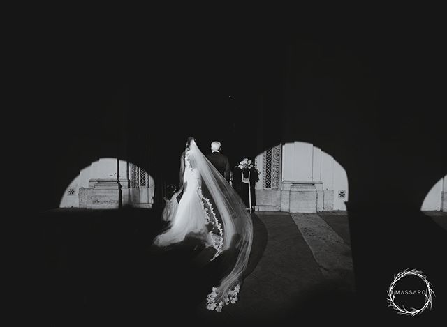 A bride and groom are walking together, the scene is monochromatic and the veil is very long. 