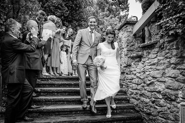 A newly married couple descends stone stairs while being celebrated by wedding guests in a black and white image.